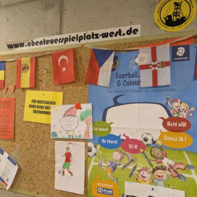 Eine Pinnwand mit verschiedenen Notizen, Fähnchen und Plakaten zu einem Fußball-Malwettbewerb. Helle Schilder und Cartoon-Bilder von Kindern, die Fußball spielen, schmücken die Pinnwand. Oben rechts befindet sich ein gelber Aufkleber mit Informationen zum Spielplatz.