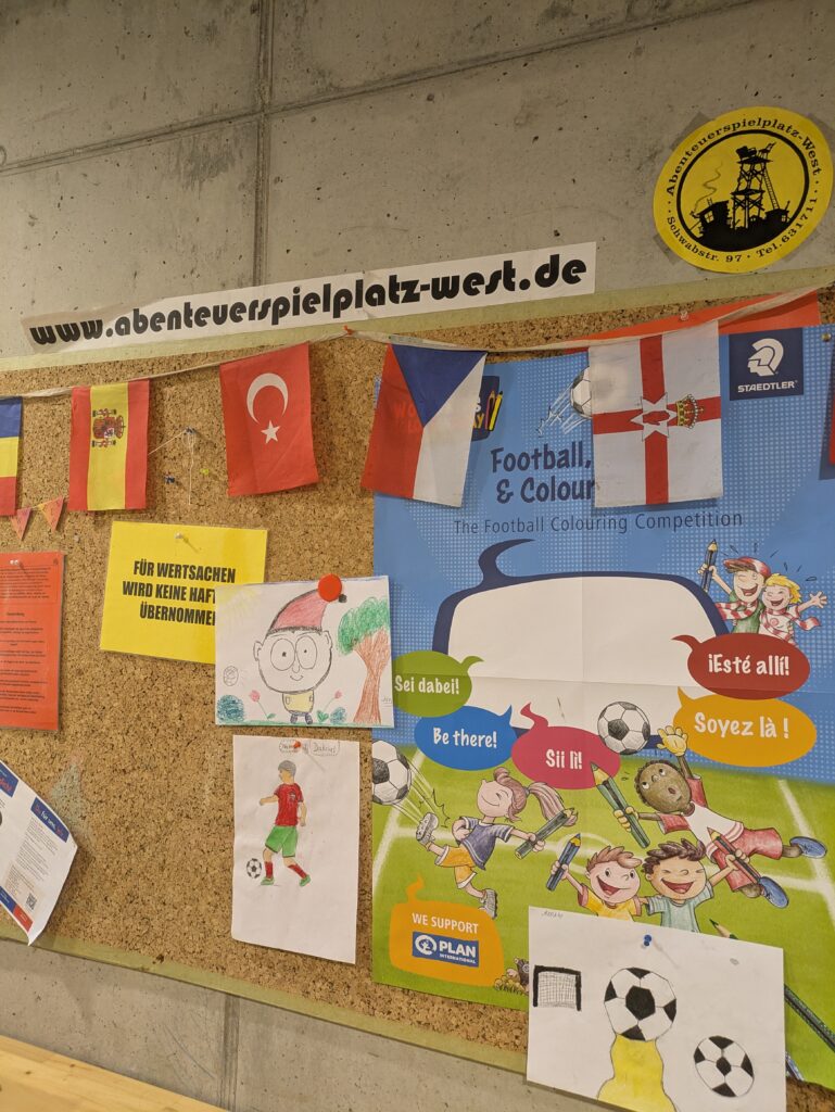 Eine Pinnwand mit verschiedenen Notizen, Fähnchen und Plakaten zu einem Fußball-Malwettbewerb. Helle Schilder und Cartoon-Bilder von Kindern, die Fußball spielen, schmücken die Pinnwand. Oben rechts befindet sich ein gelber Aufkleber mit Informationen zum Spielplatz.